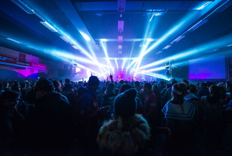 Photo taken from the back of a busy club, looking at the stage, with blue and purple lights