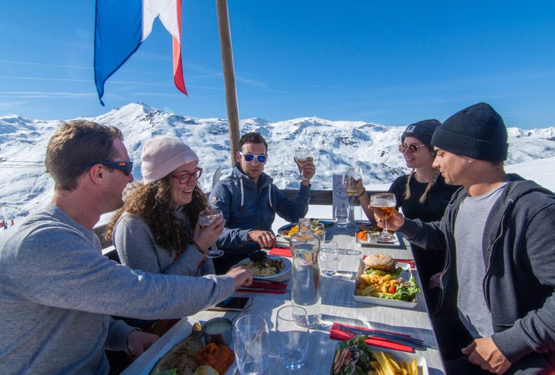 A group of friends sit outside on a mountain restaurant terrace with drinks and food, on a blue sky day