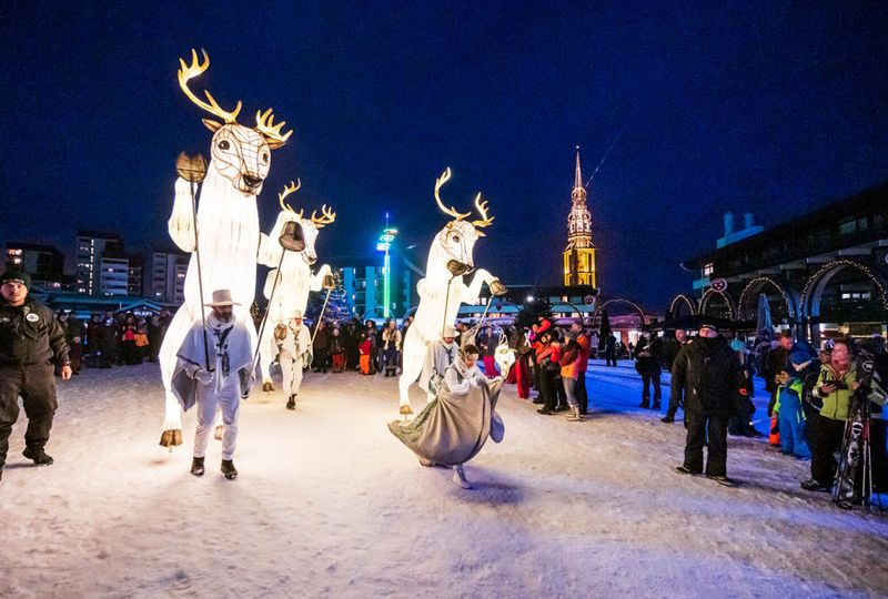 Entertainers dressed up as stags and covered in lights, parade in front of crowd outside on the snow at night time. A lit up church is visible in the background