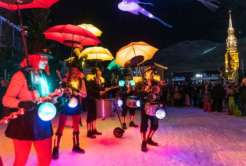 A night show act, with musicians under colourful umbrellas attached to them, play on snow under colourful lights under a dark sky