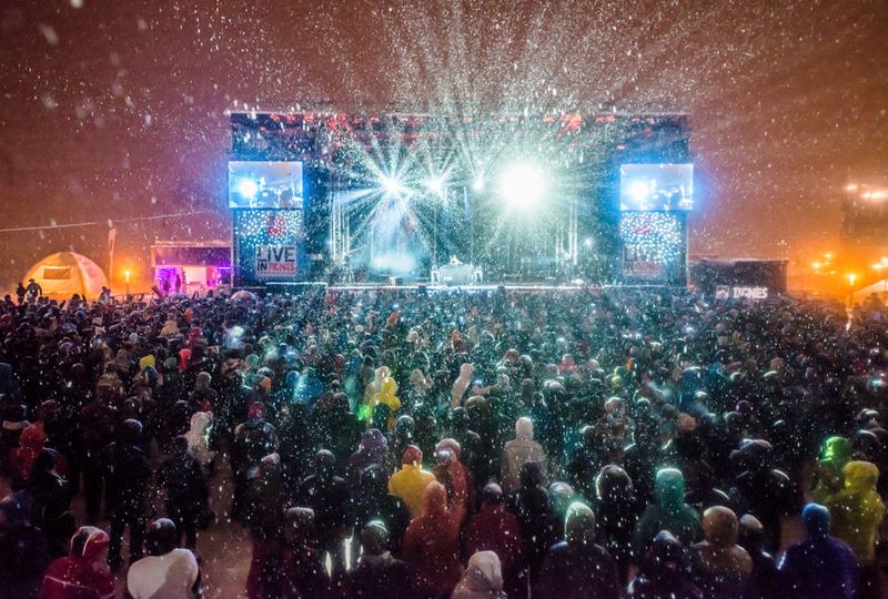 Snow falls as a crowd watches a band on stage at night time, during a concert or festival
