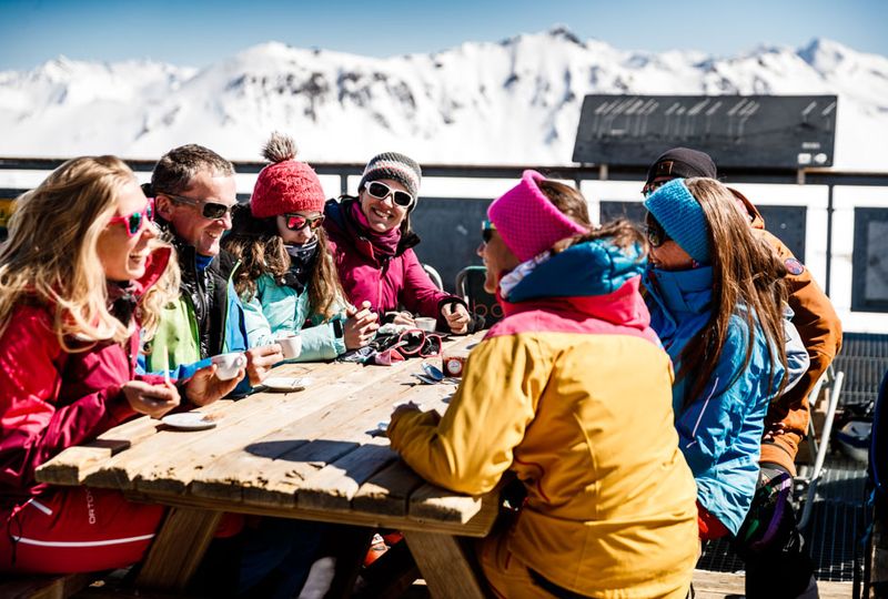 six friends sit around a picnic table drinking espresso wearing sunglasses and hats, on a coffee break from skiing
