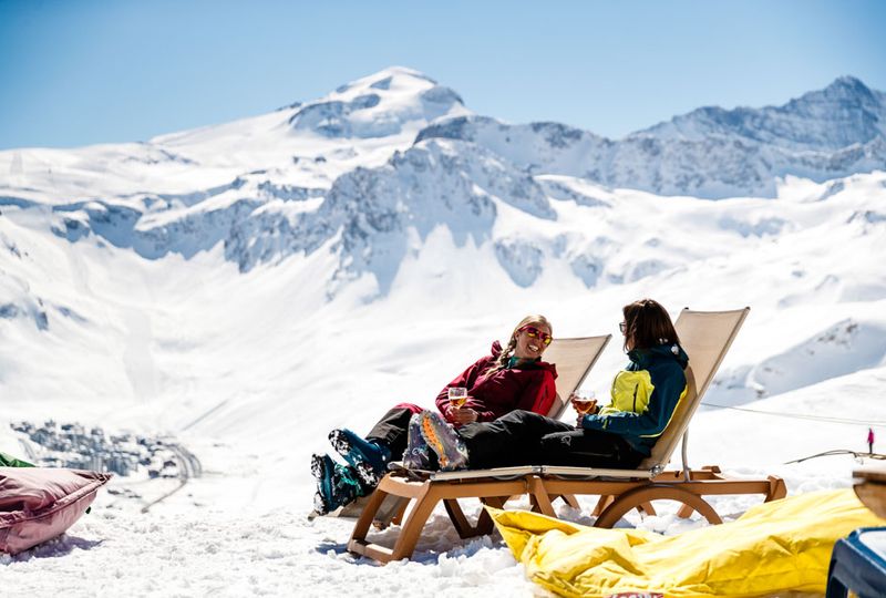 Two skiers take time out on sun loungers  on the top of a mountain, sipping wine as they chat