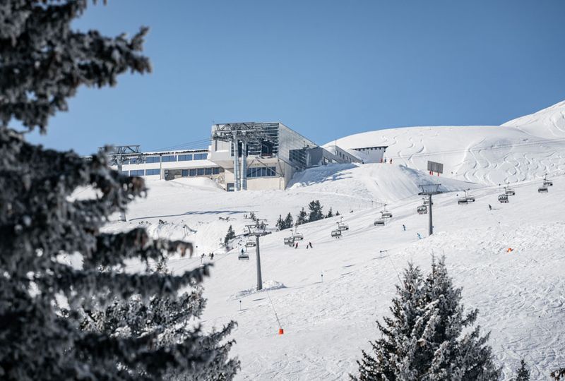 Image taken through the trees of a top lift station, very modern in look with reflective mirror-like glass. A chair lift sits in the mid-ground while skiers ski on the run underneath