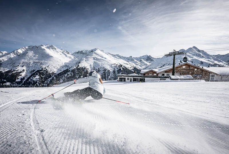 A skier in white carving the groomed slopes in St Anton, a restaurant building and gondola visible in the background