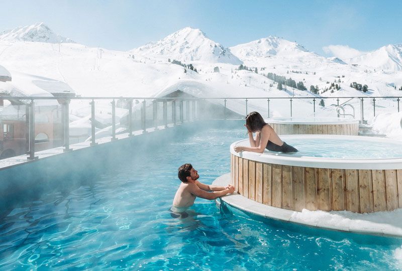 Two adults sit in a hot tub and pool, chatting, in a snowy landscape