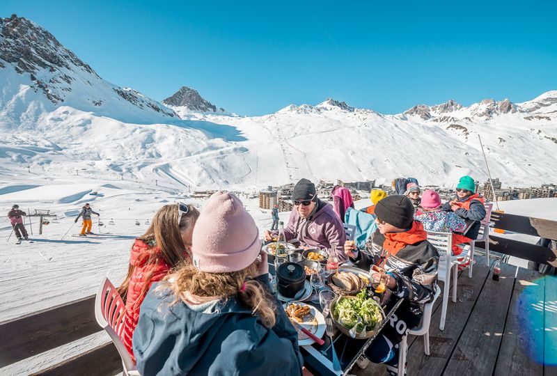 Four sit around a table on a mountain terrace at lunch time. The table is full of food, the ski slopes are visible behind