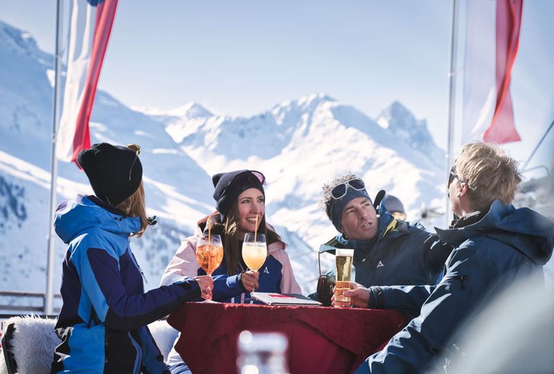 four friends sit around a table chatting with cocktails., out on the mountain with high white snowy peaks visible in the distance behind