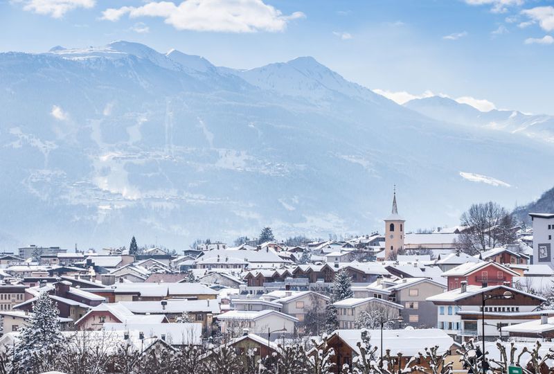 An alpine village covered in snow, the spire of a church rising high above all else except the high mountains that back the village in this shot