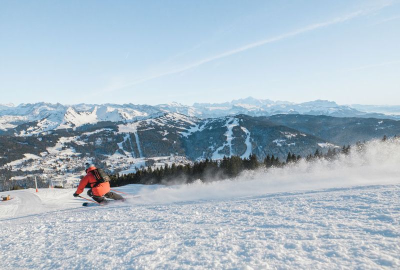 A skier in red carves down a gentle slope, the mountains with ski runs cut into the alpine visible in the distance ahead
