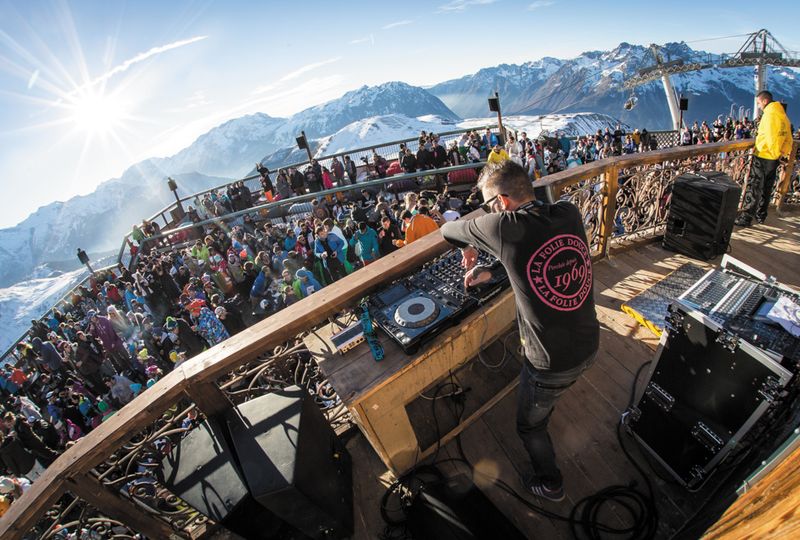Party skiers enjoy a day-time party on a sunny mountain deck, in front of a DJ