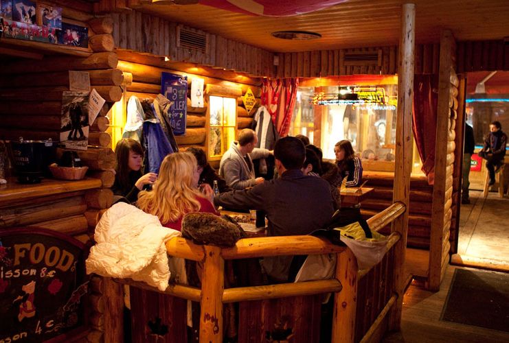 A cosy indoor mountain bar is shot at night time, warm lighting and friends around a table - most of the inside of the cabin is made of wood
